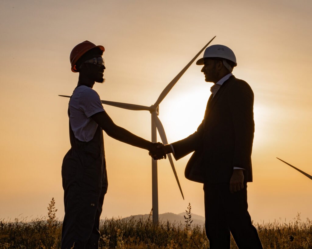 Silhouette of african american engineer and indian inspector using digital tablet during meeting outdoors and shaking hands. Two partners standing on field with wind turbines during amazing sunset.